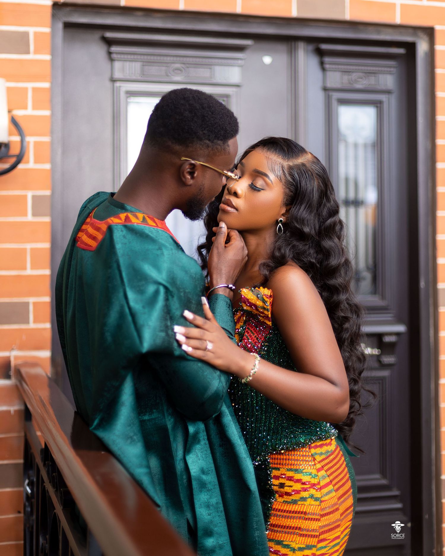 A couple shares a romantic moment in their traditional formal attire against a brick wall and black door.