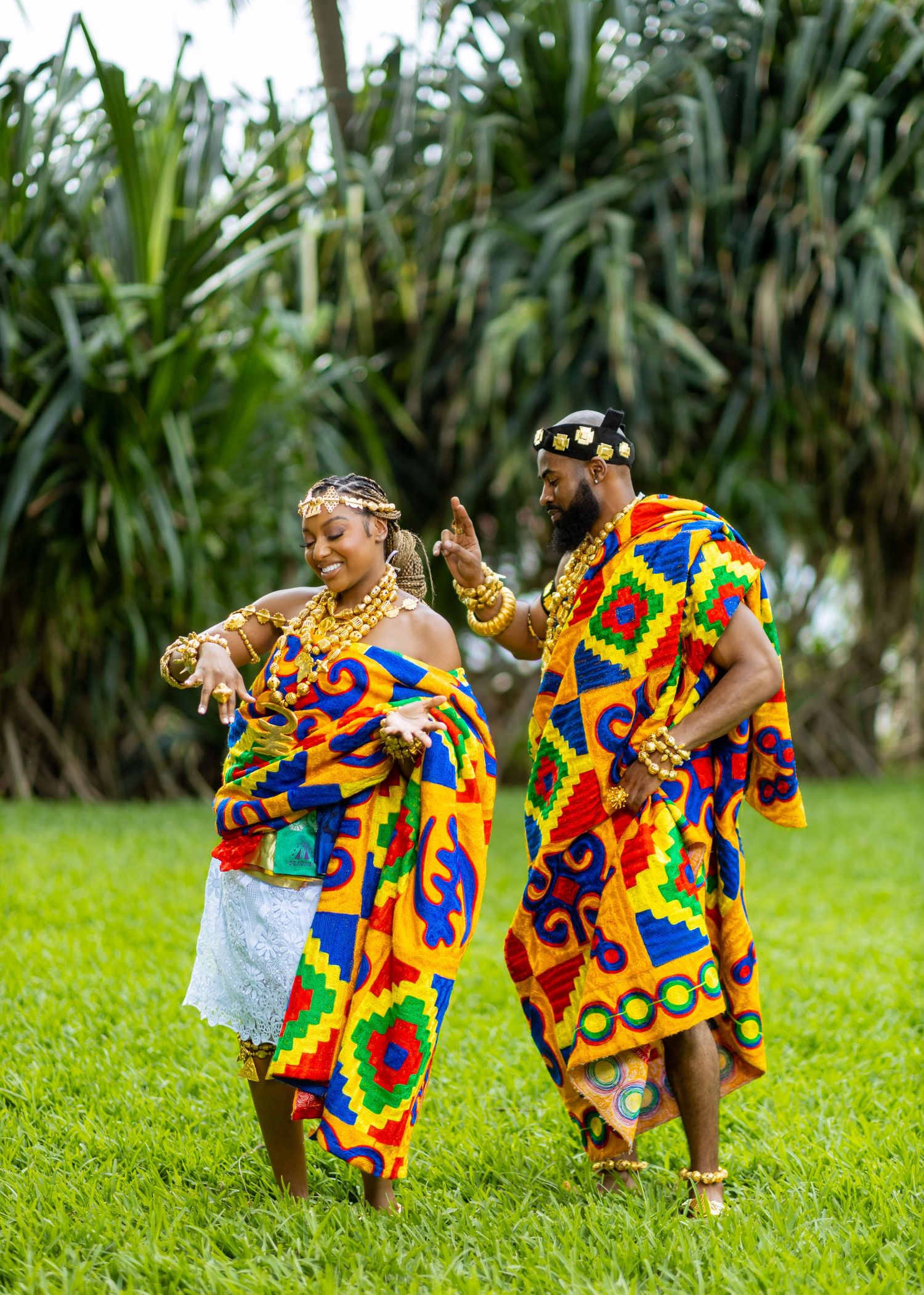 Children in vibrant African kente cloth dance joyfully together on a grassy field by palm trees.