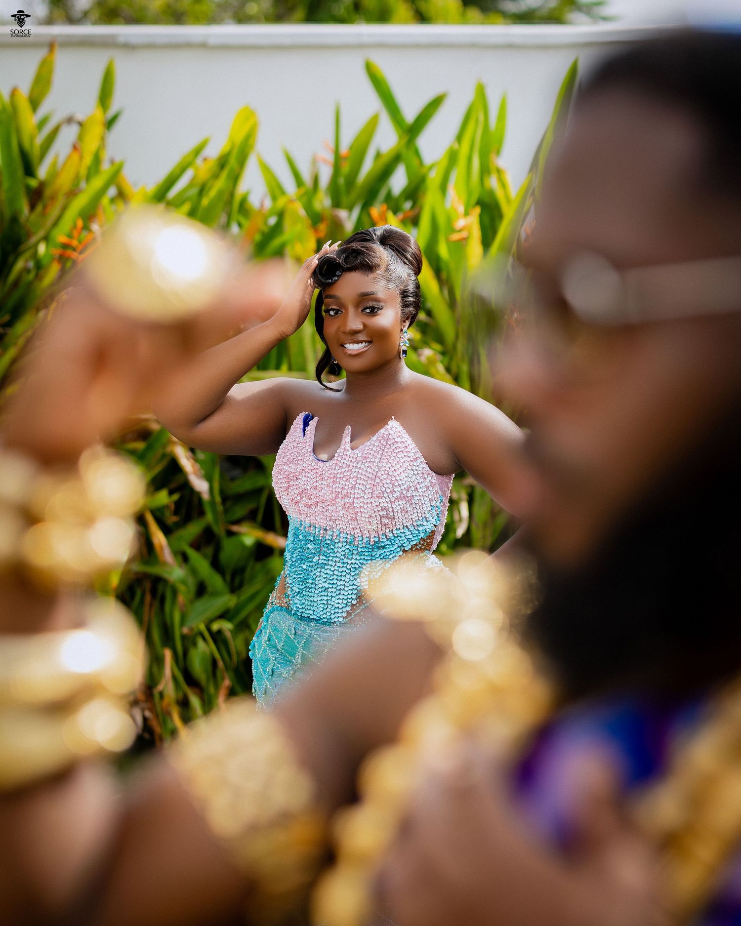A woman in a beaded teal and pink dress poses outdoors while a man in gold jewelry stands in the foreground.