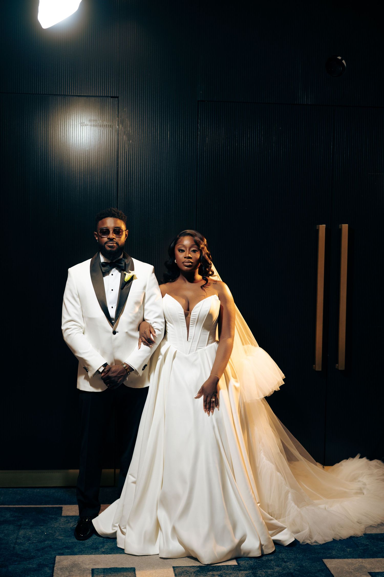 Elegant couple poses in formal wedding attire with a white tuxedo and flowing bridal gown against a dark backdrop.