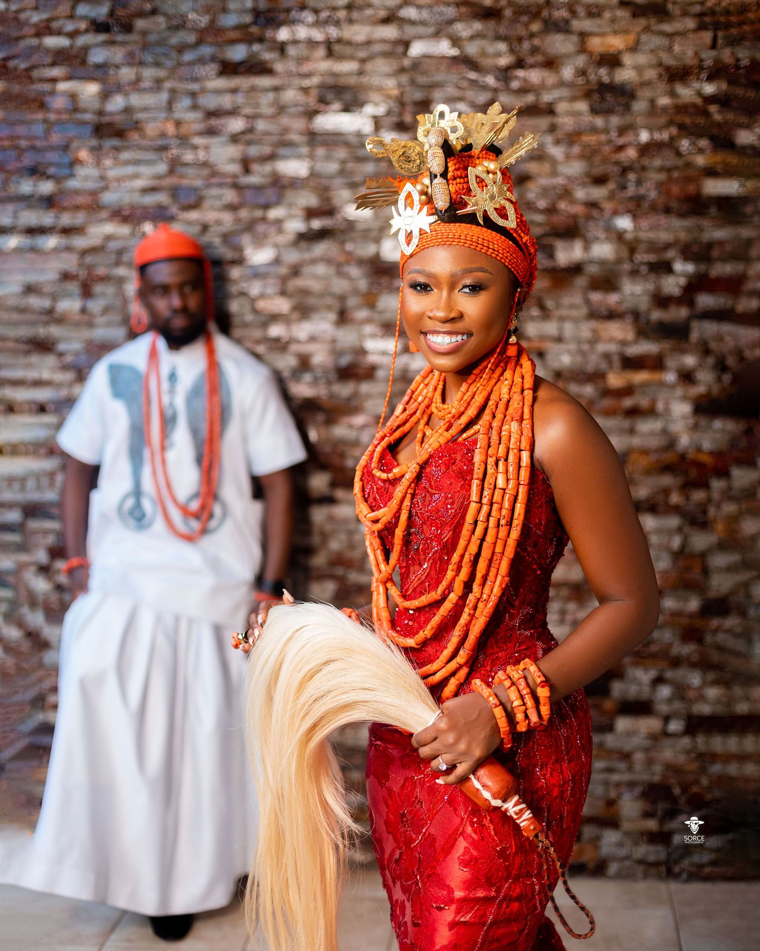 Traditional Nigerian bride wearing ornate coral beads and a red dress poses against a rustic brick wall.