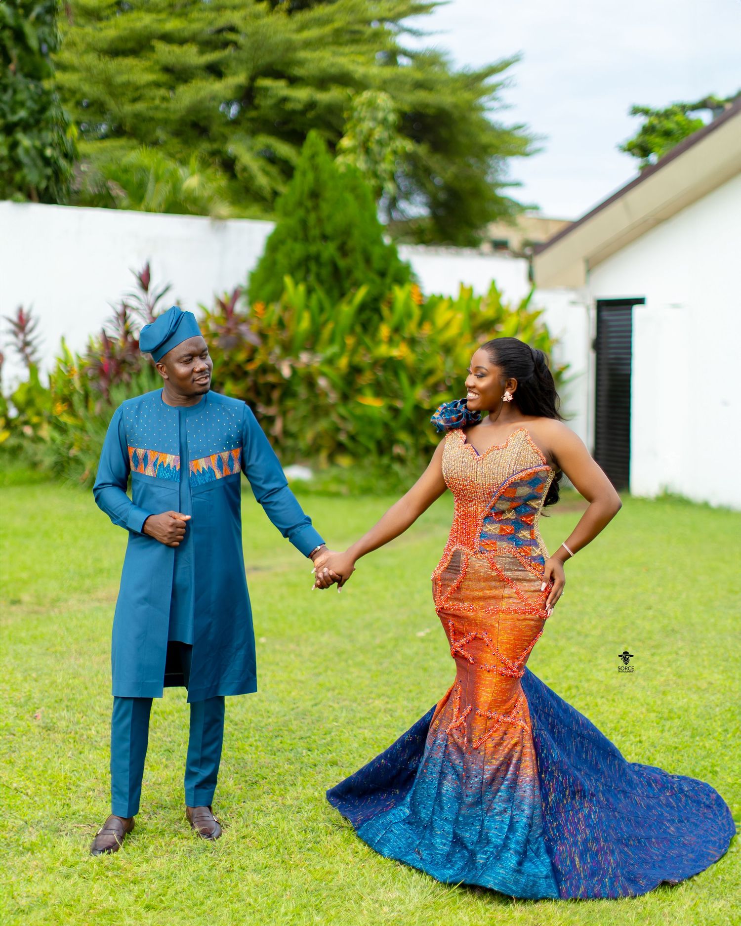 Couple in coordinating blue and orange African print outfits dancing playfully on a green lawn.