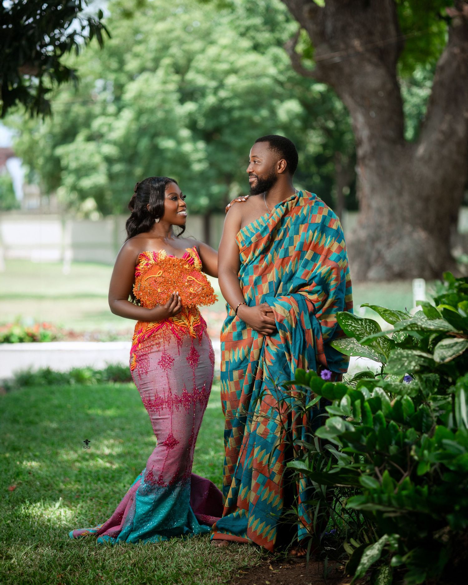 Two people dressed in vibrant African-inspired formal attire share a joyful moment together in a lush garden setting.