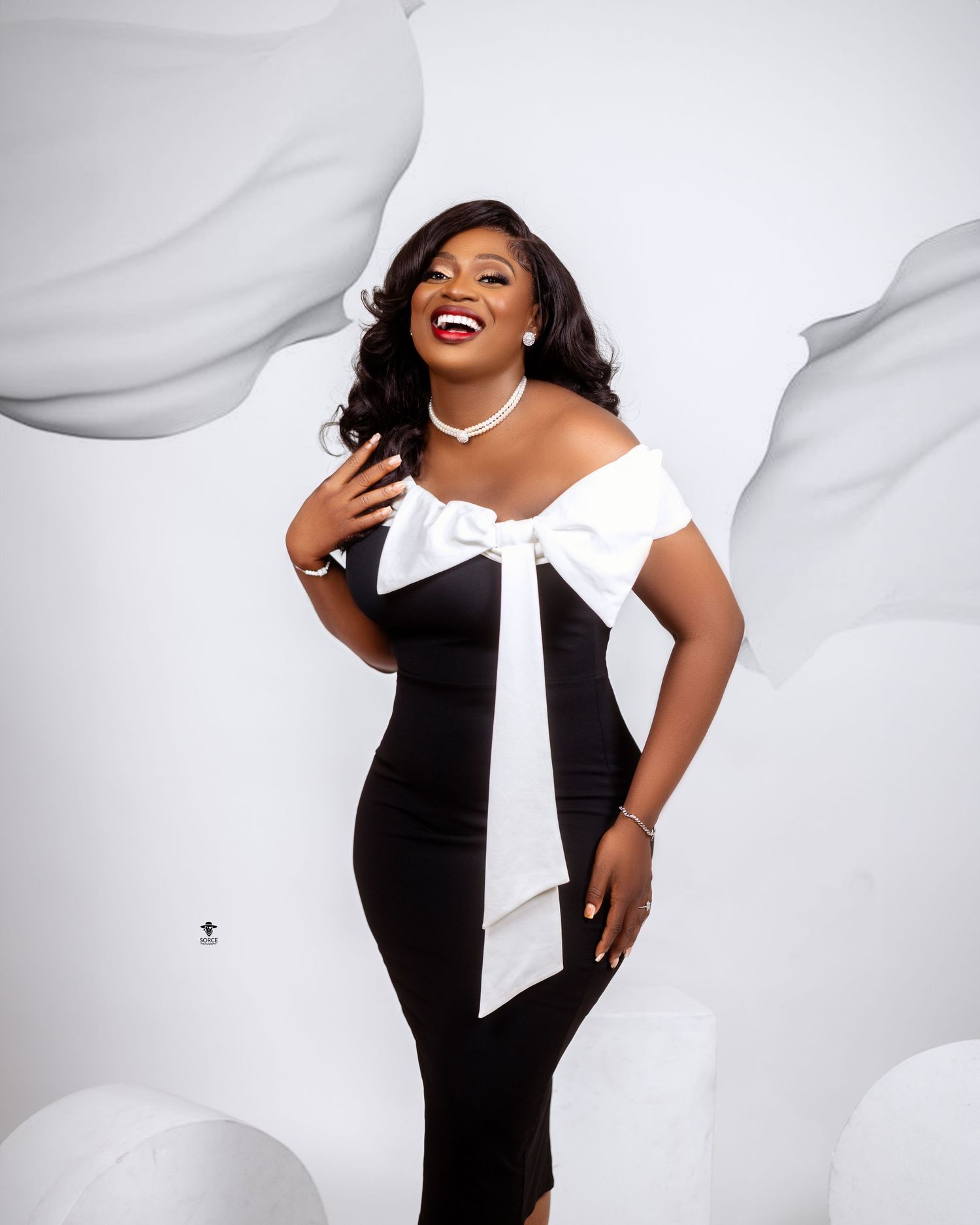 Figure in a black and white dress with dramatic large bow poses joyfully against a white studio backdrop.