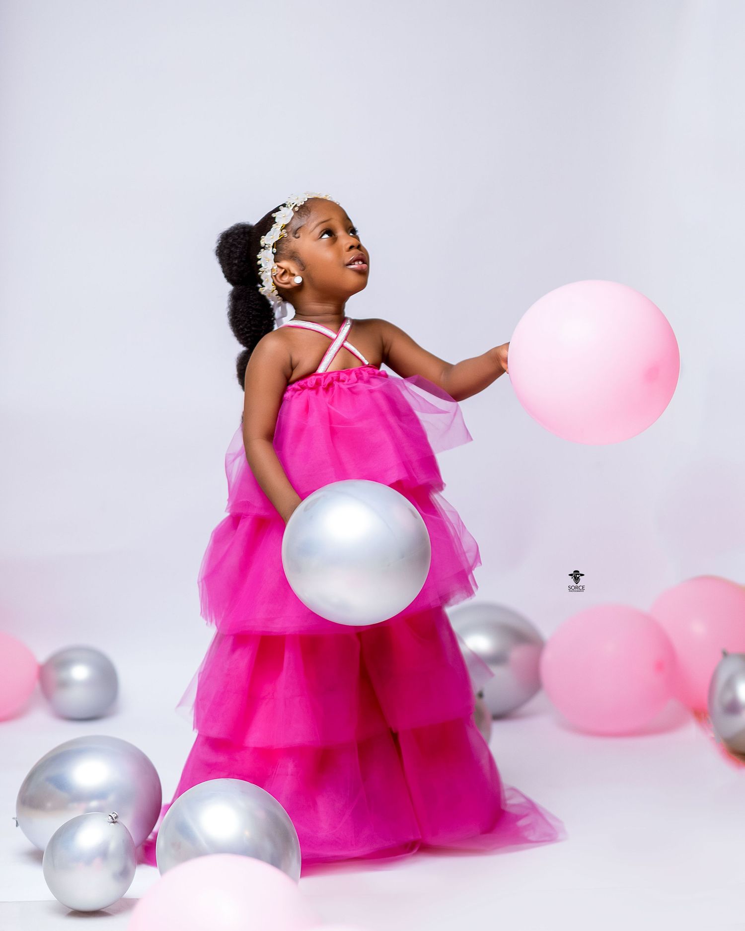 A young girl in a bright pink dress playfully poses with pink and silver balloons against a white backdrop.