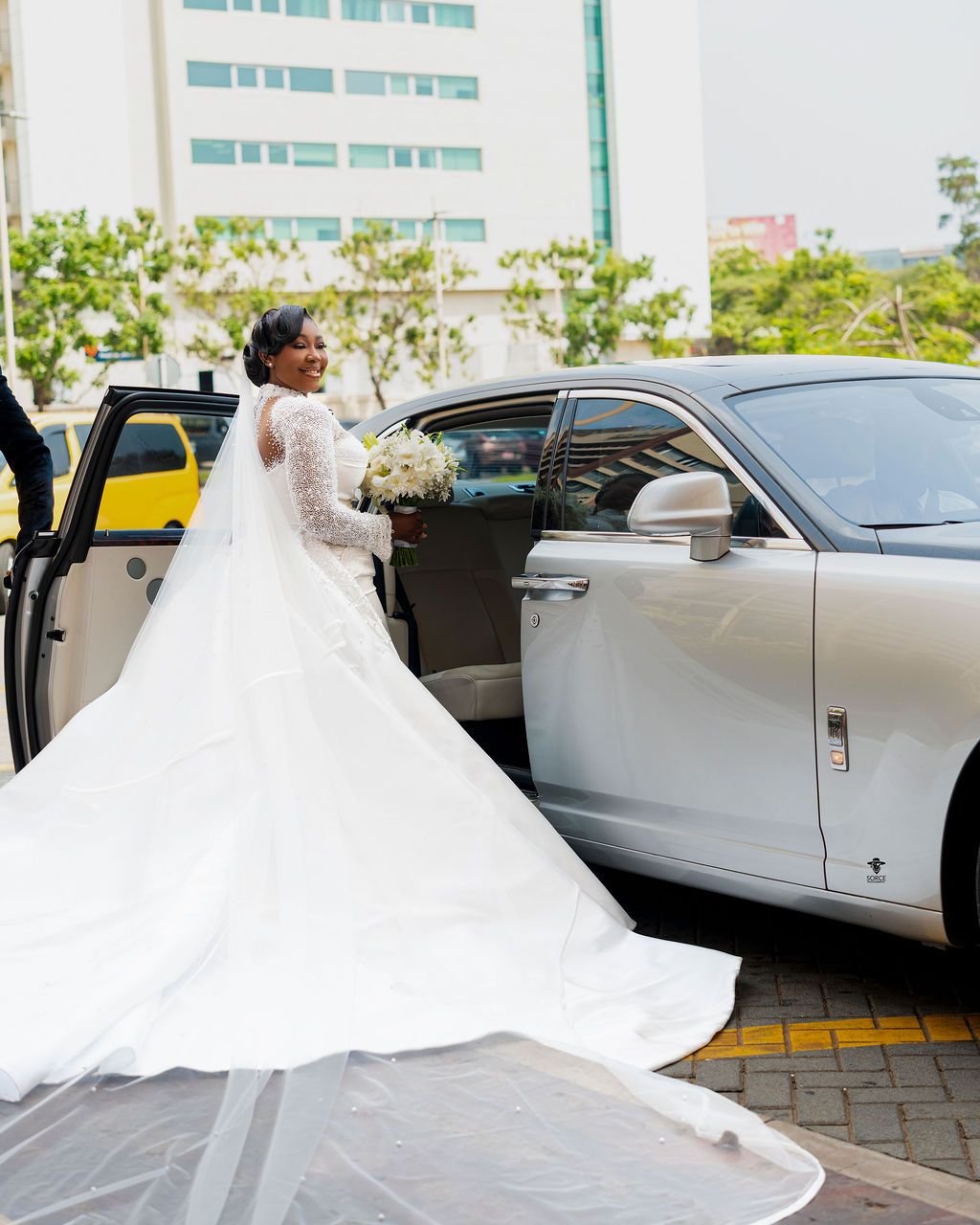 Bride in elegant long-sleeve white gown with cathedral train stands beside luxury white car holding bouquet.