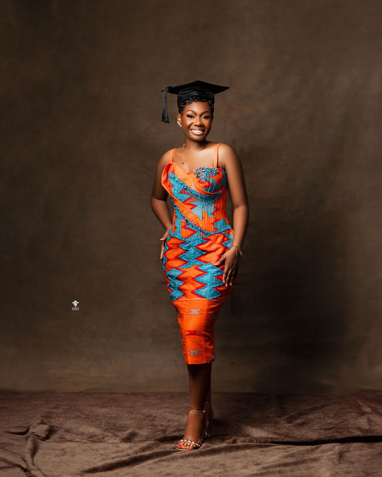 Graduate wearing vibrant orange and blue patterned dress with graduation cap poses in a professional studio portrait.