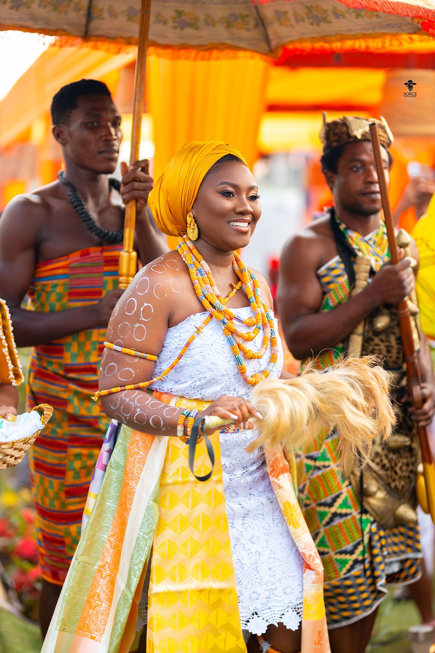 Colorful traditional African wedding celebration with dancers wearing vibrant kente cloth and white lace attire.