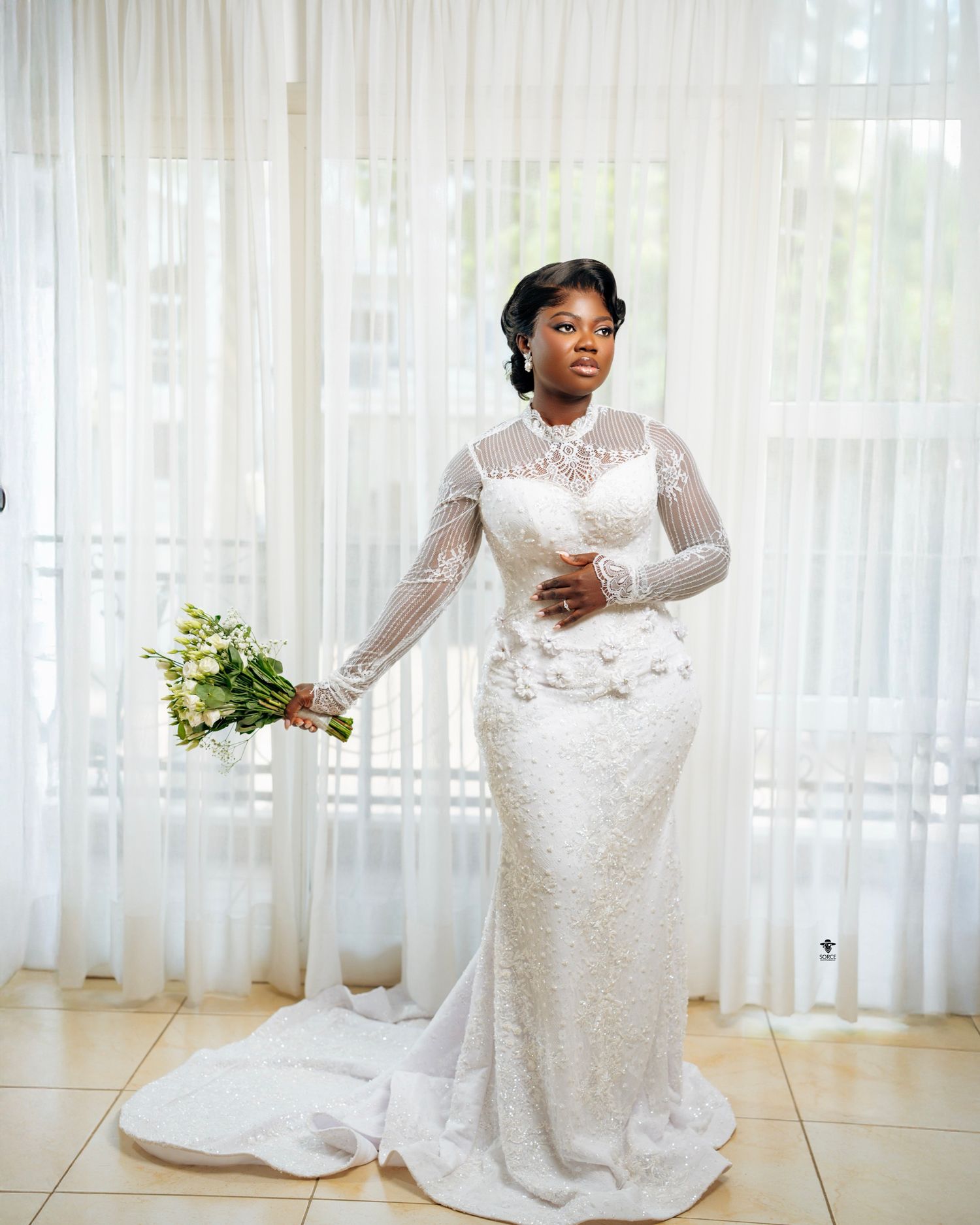 Elegant bride in long-sleeve lace wedding gown holding bouquet, standing by sheer white curtains in natural light.