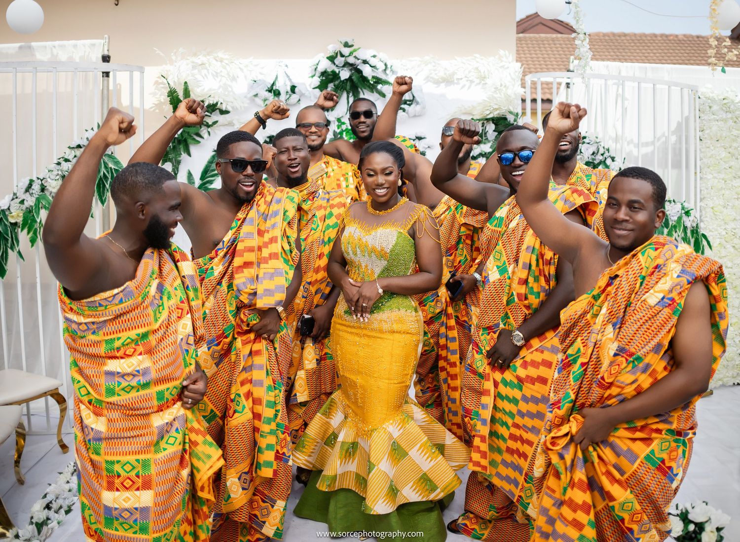 Wedding guests in vibrant yellow and green kente cloth dance and celebrate during a traditional African ceremony.