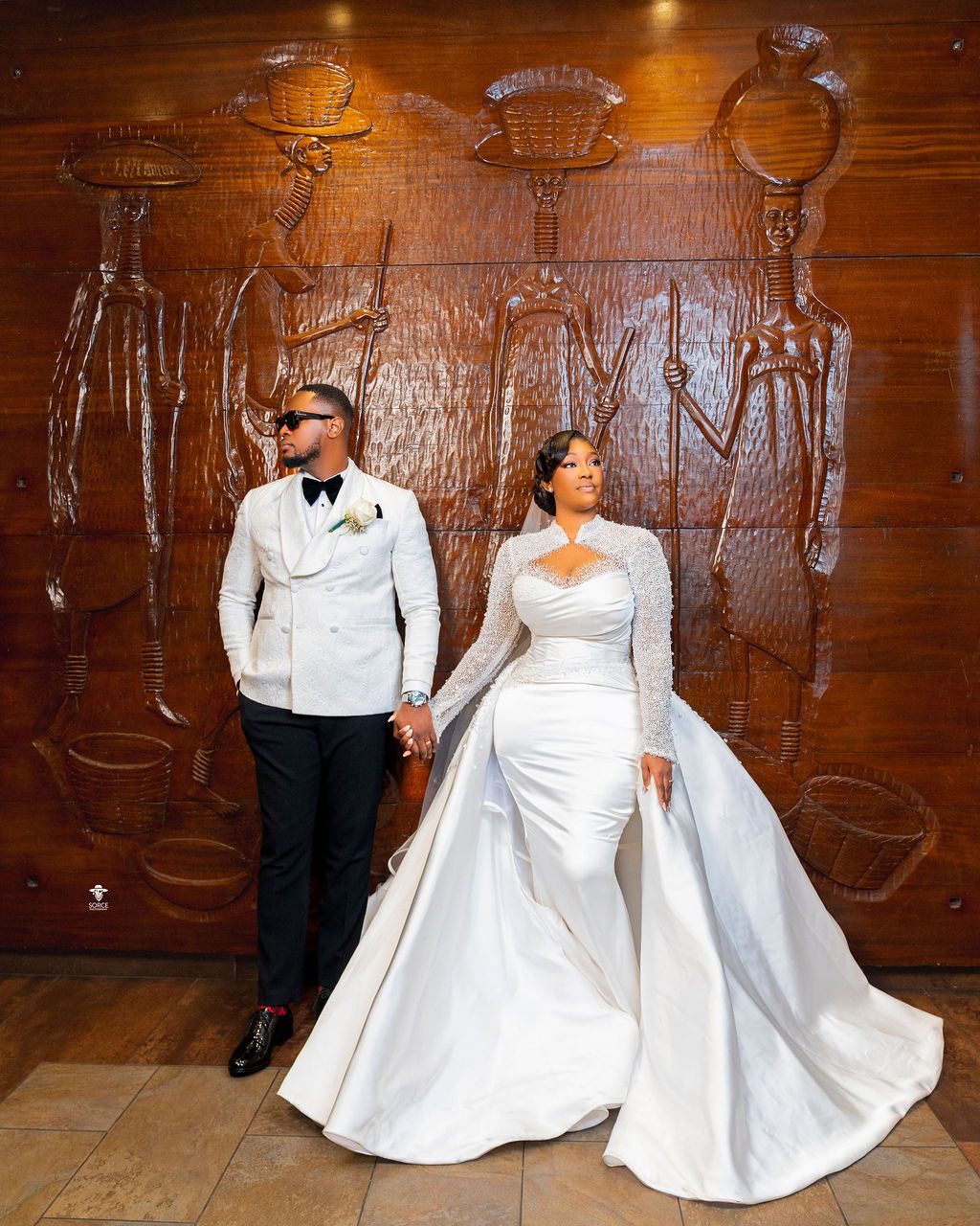 Elegant couple in white wedding attire posing in front of an African-inspired wooden carved wall mural.