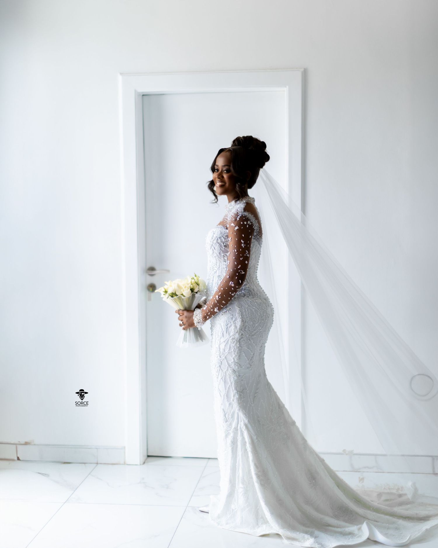 A bride in a long sleeve lace wedding dress poses against a minimalist white background in a studio setting.