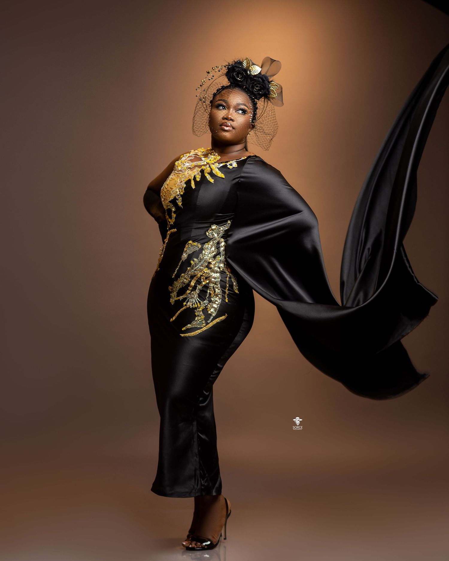 Fashion model wearing black flowing dress with gold embellishments poses dramatically against brown studio backdrop.