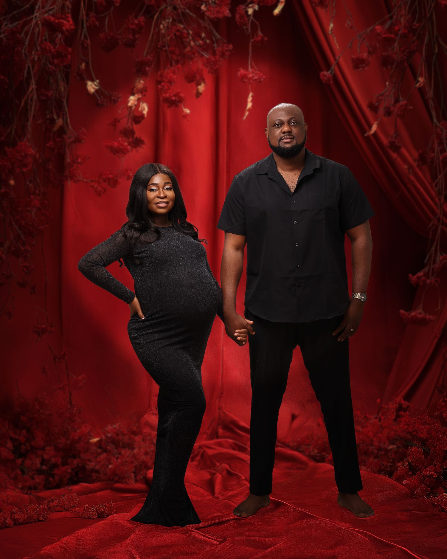 A maternity photo shoot against a deep red backdrop featuring two people in black outfits holding hands.
