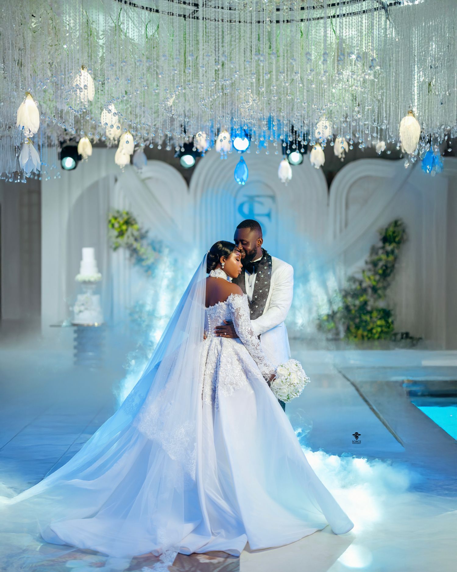 A bride and groom share a romantic embrace on a dreamy wedding dance floor surrounded by crystal chandeliers and soft blue lighting.