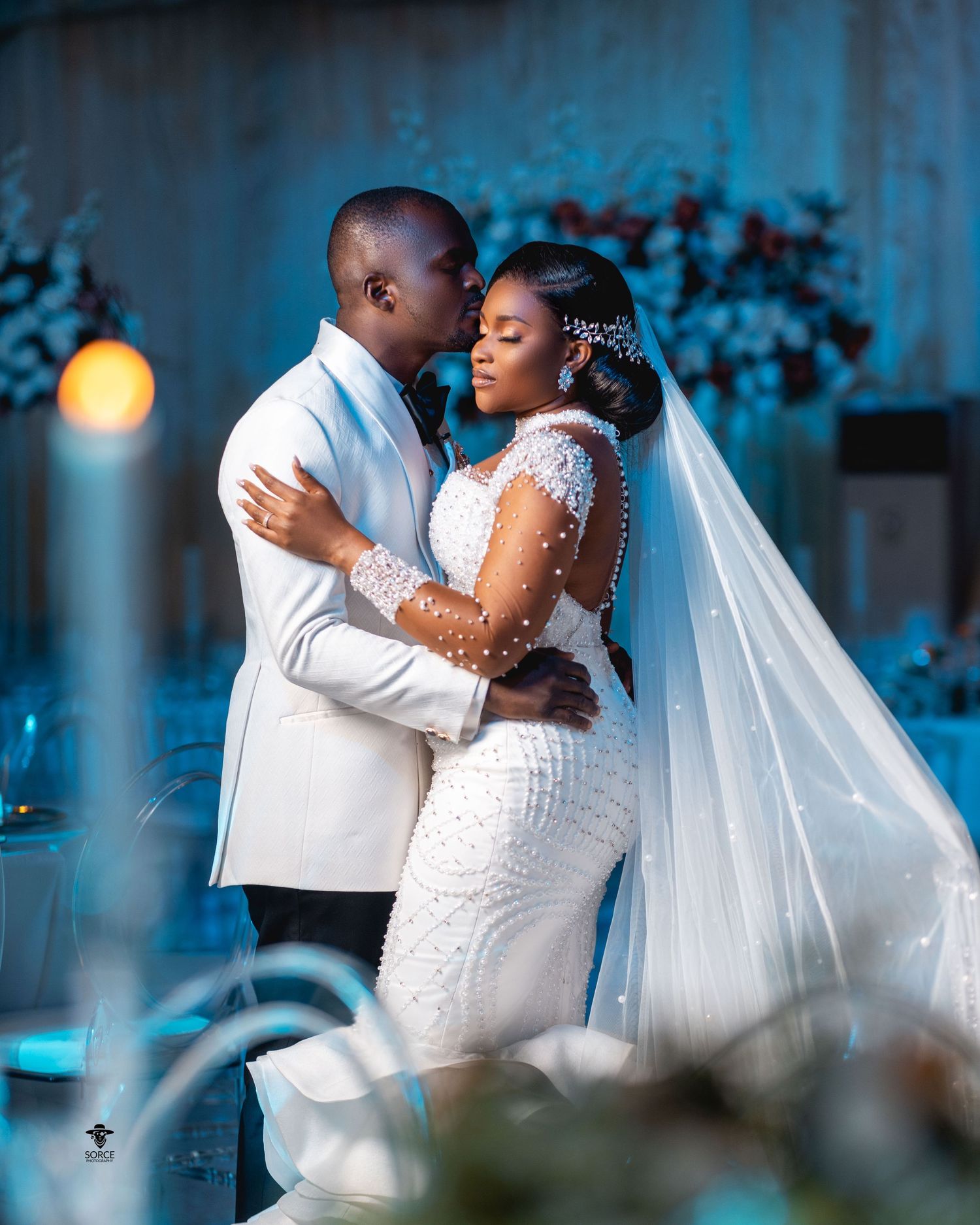 A couple embraces during their wedding ceremony with blue-tinted lighting and floral decorations in the background.