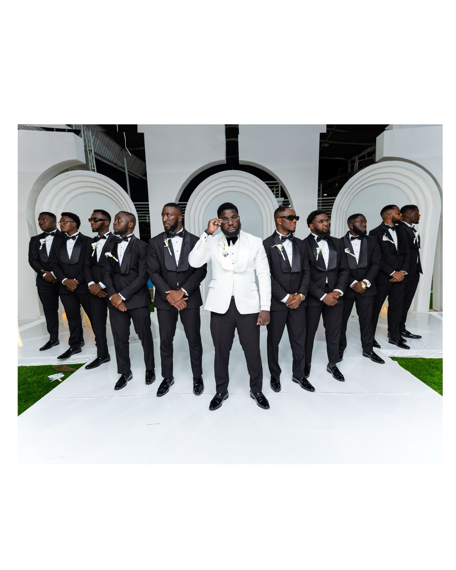 Groom in white tuxedo jacket poses with groomsmen in black tuxedos against white arch backdrop.