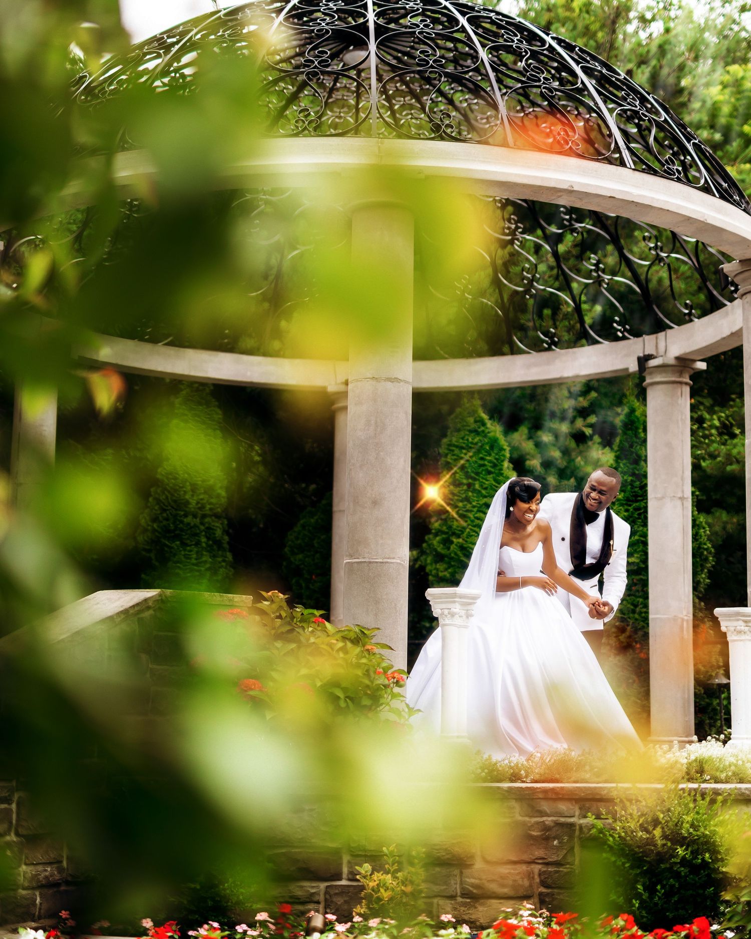 A couple sits romantically under a garden gazebo surrounded by lush greenery and red flowers during an outdoor celebration.