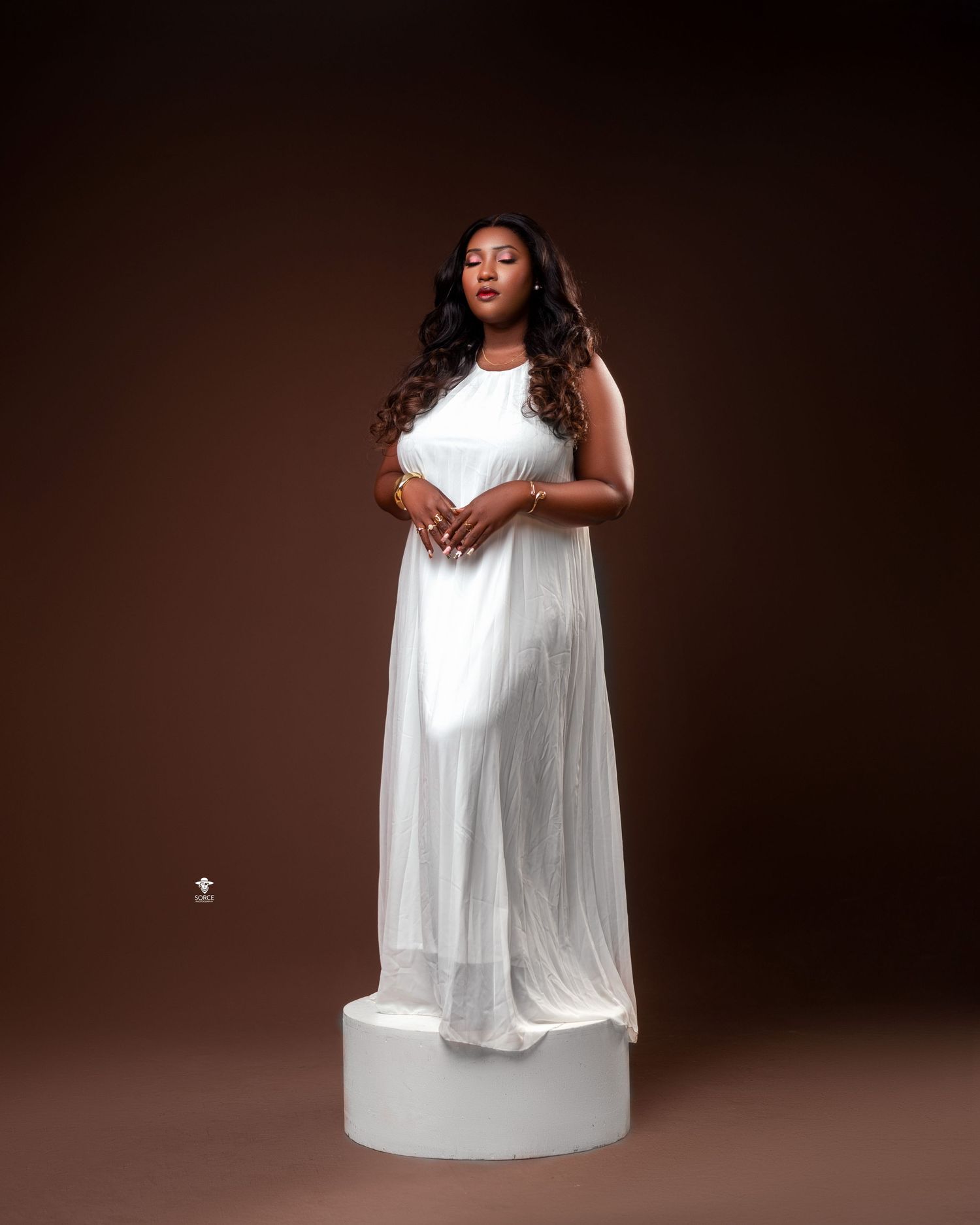 Model poses elegantly in flowing white dress against dark brown backdrop in professional studio photoshoot.