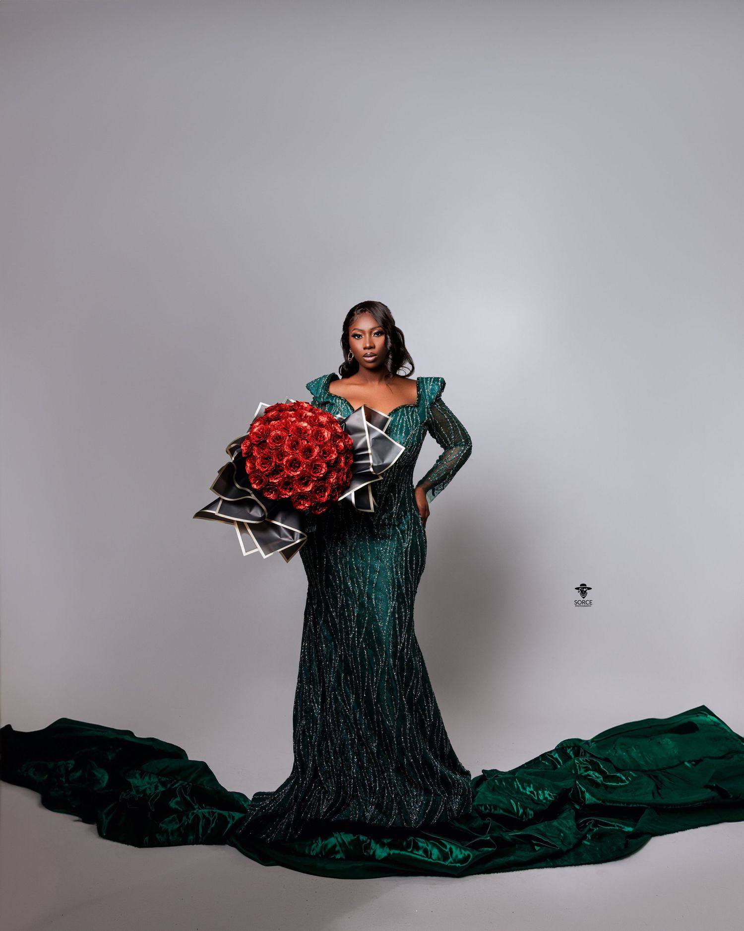 Model wearing elegant emerald green textured gown with long train holding large red rose bouquet against gray backdrop.