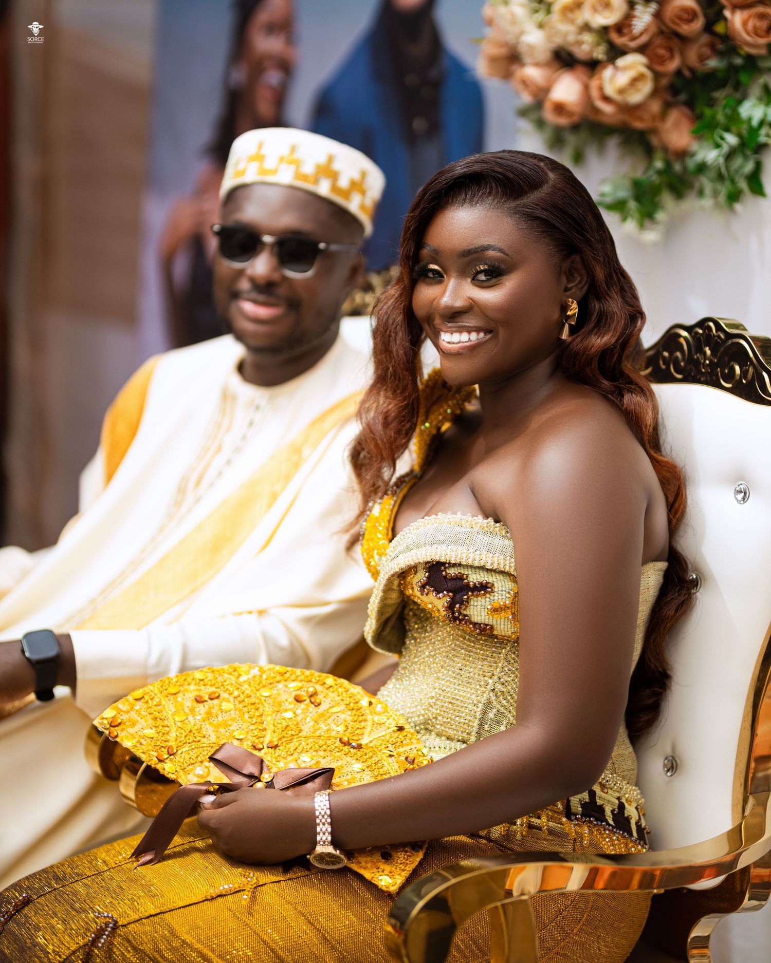 Wedding guests in traditional Ghanaian attire seated at reception tables with gold decor and floral arrangements.