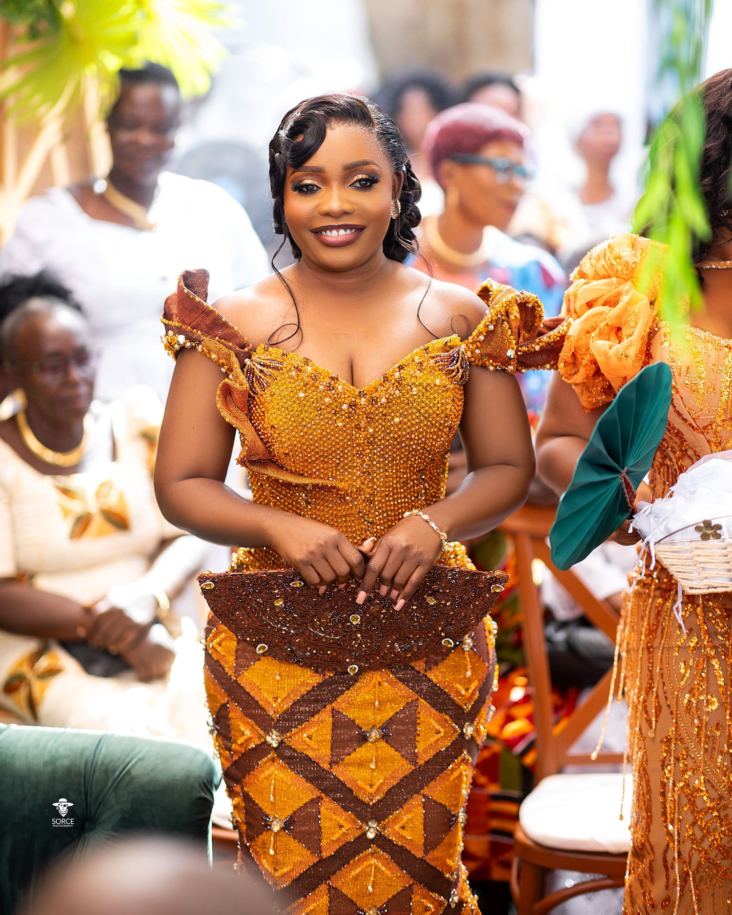 Traditional West African celebration featuring guests wearing vibrant orange and gold kente print dresses with beaded details.