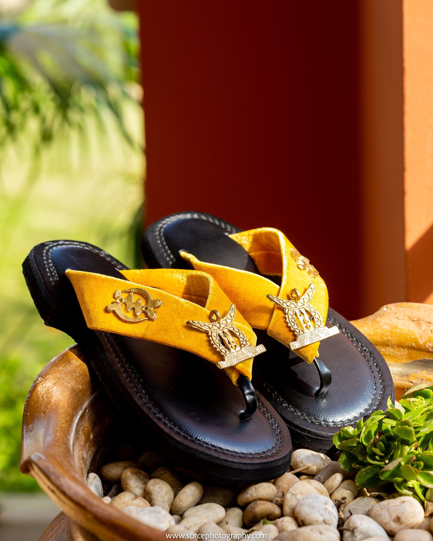 Yellow and black leather thong sandals with gold angel emblem decorations displayed on pebbles in a wooden bowl.