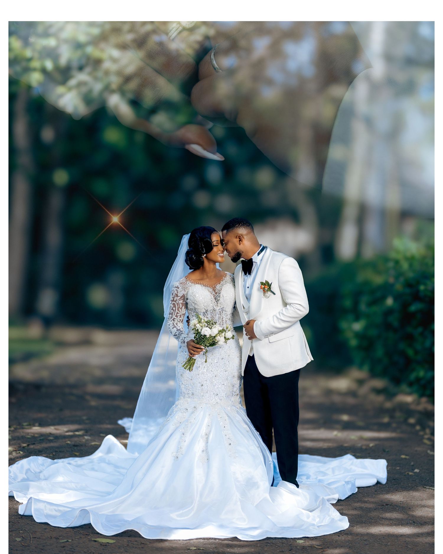 Elegant wedding photo on a tree-lined path showing a couple in white formal attire with a flowing lace gown and bouquet.
