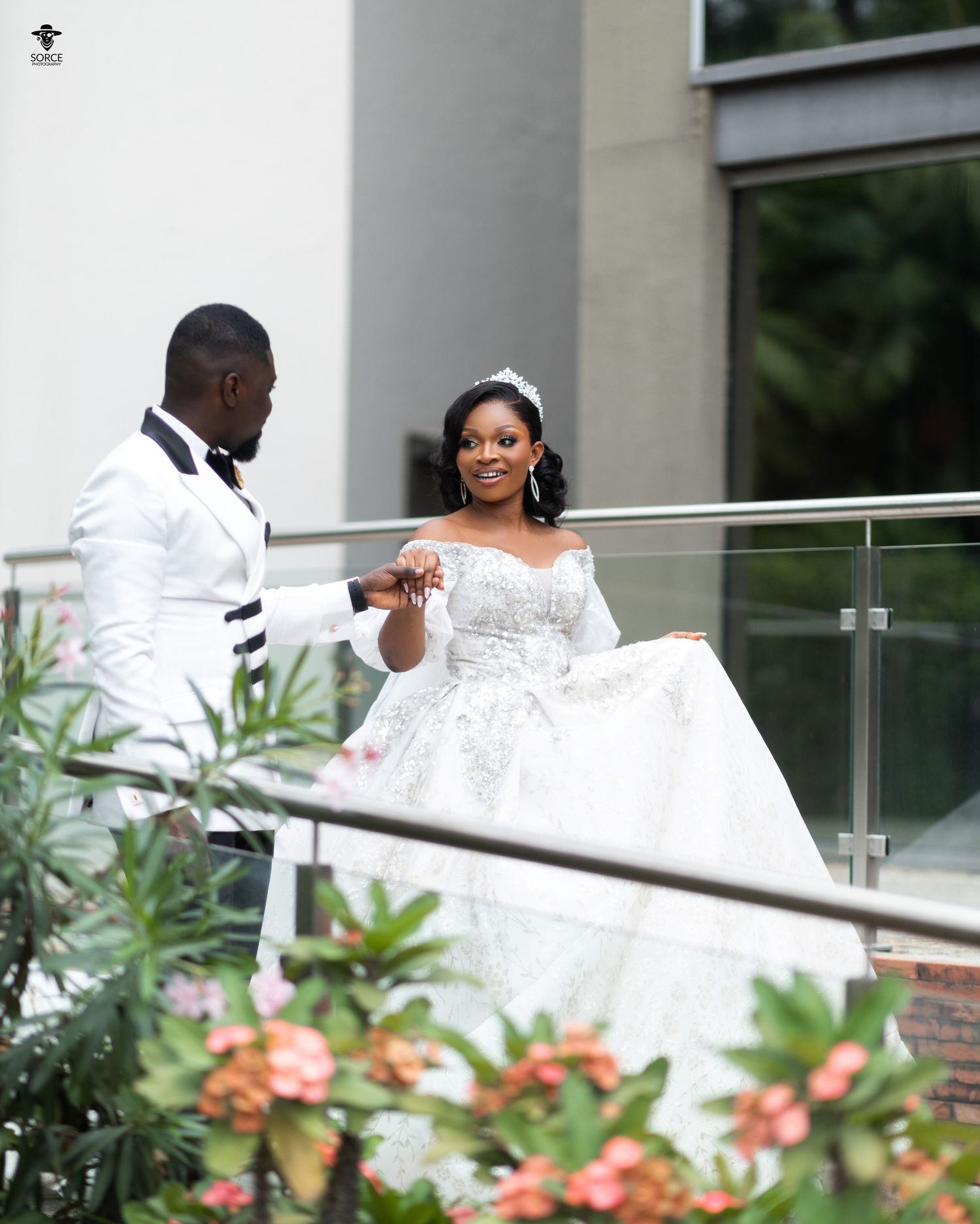 A couple in white wedding attire stand together on an outdoor balcony with flowering plants in the foreground.