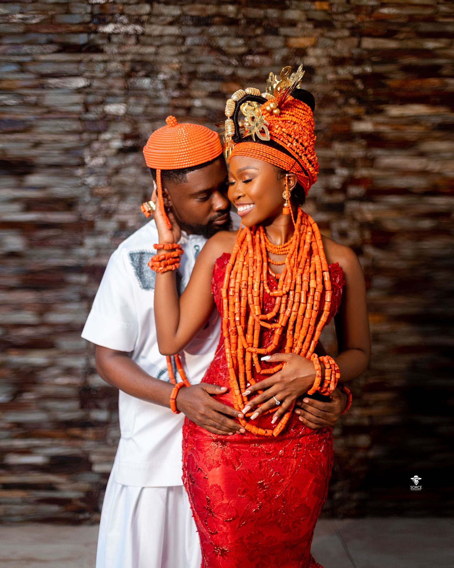 A traditional Nigerian wedding couple embraces while wearing ceremonial coral beads and red attire against a rustic wall.