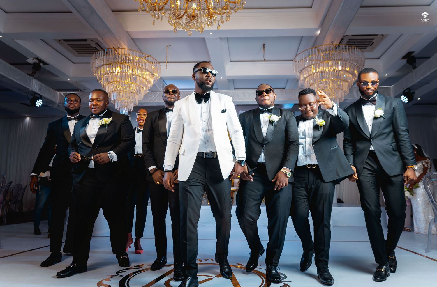 Wedding party in black tuxedos and white jackets dancing in a line at a formal reception with crystal chandeliers.
