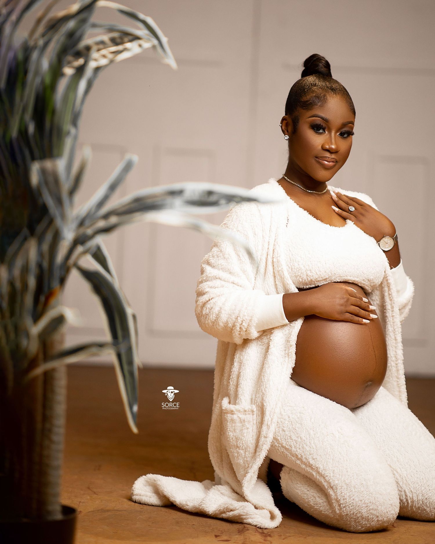 An expectant mother in white loungewear and cardigan poses elegantly in a maternity photoshoot with indoor plants.