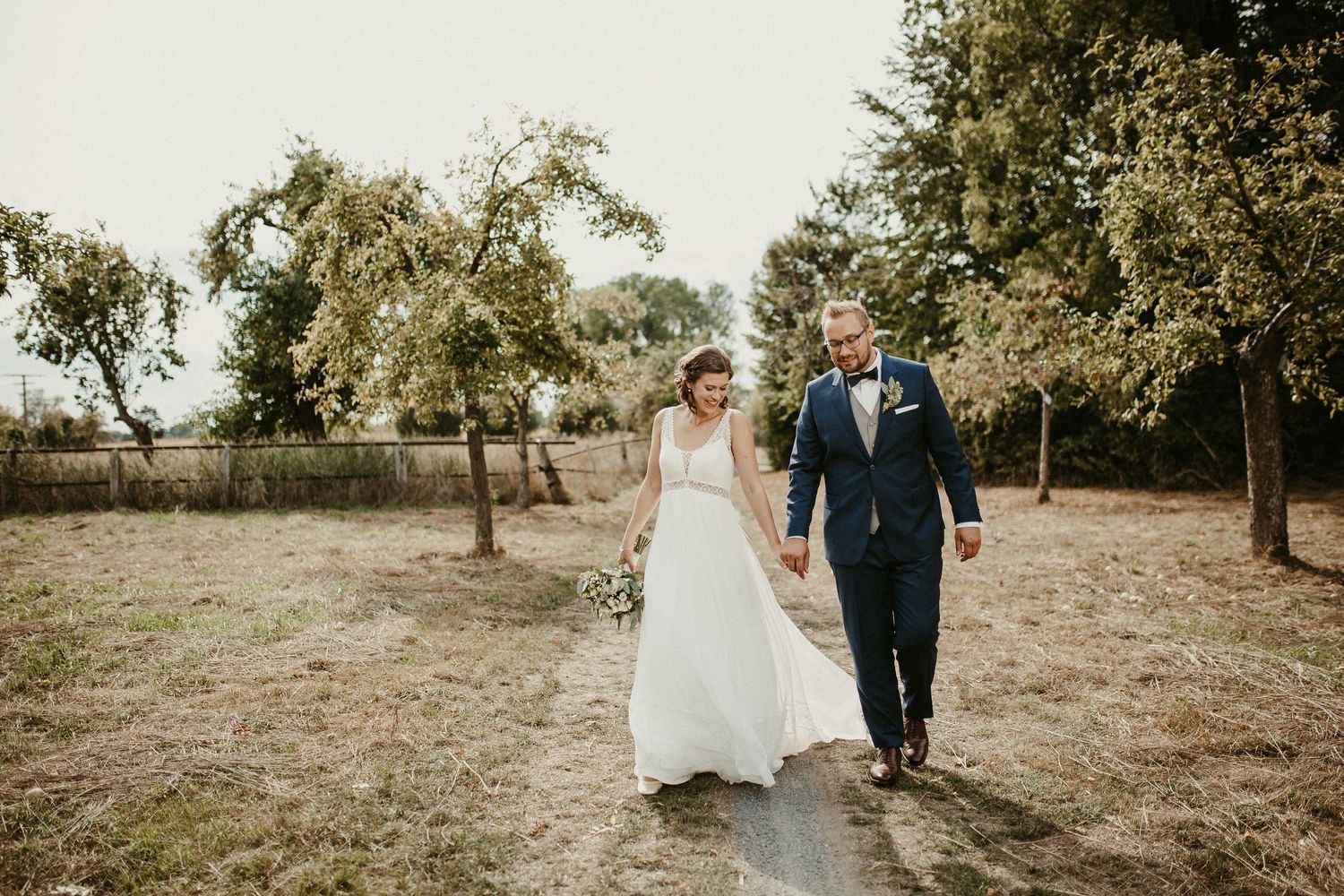 Jungvermählte gehen an ihrem Hochzeitstag Hand in Hand durch einen Obstgarten.