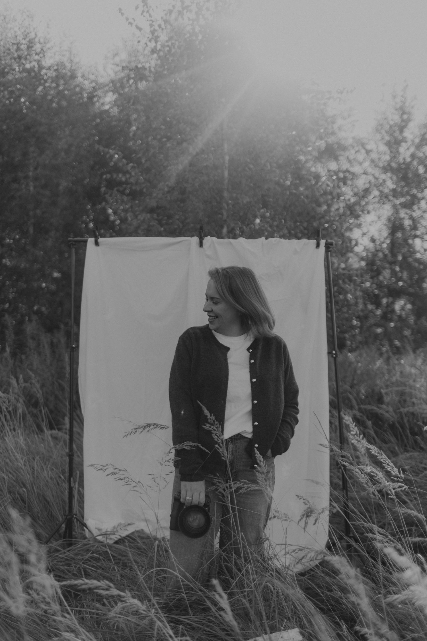 Black and white artistic portrait in tall grass with white backdrop showing figure in dark jacket against natural setting.