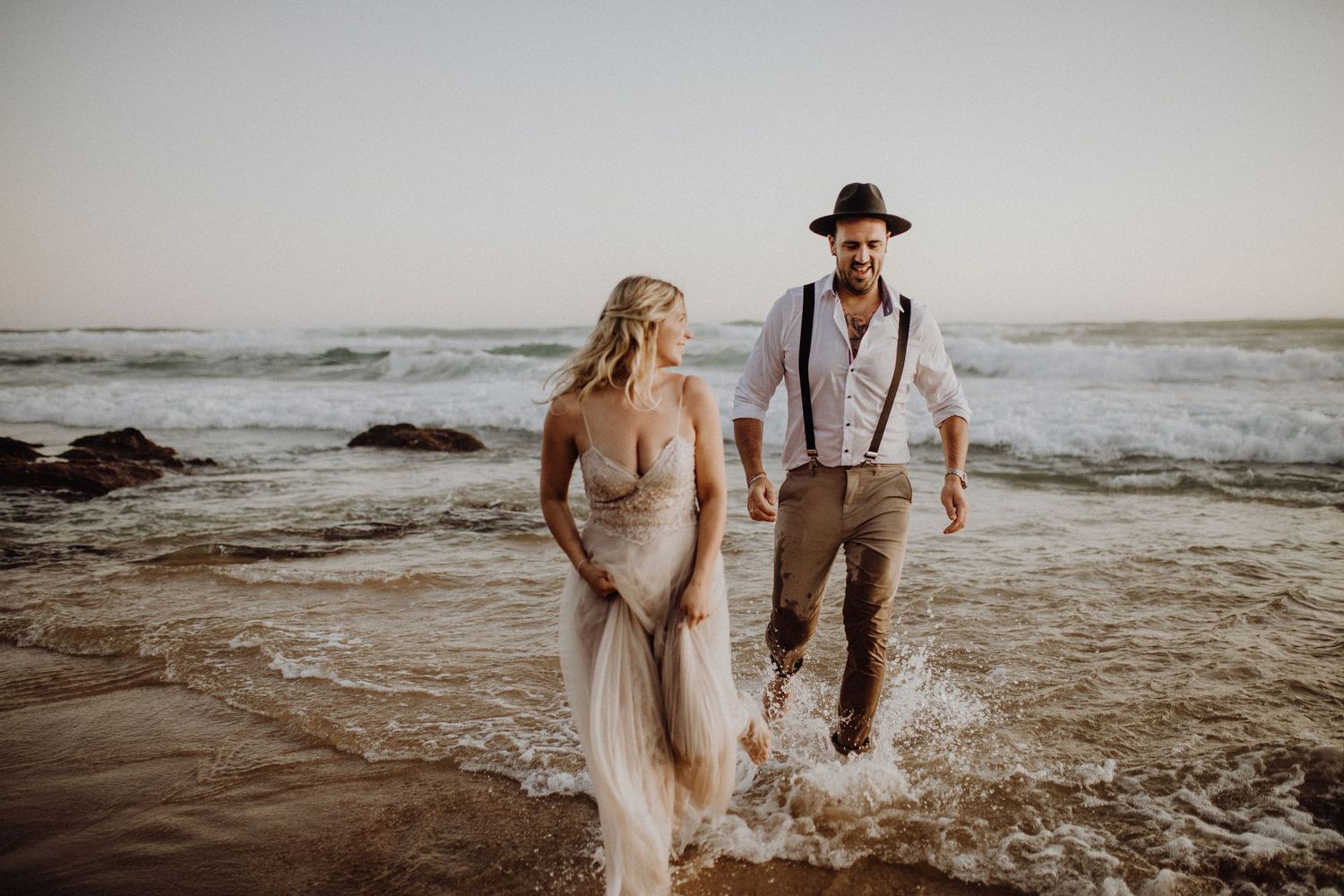 Romantisches Strandfotoshooting mit einem Paar, das bei Sonnenuntergang durch die Meereswellen watet in Südafrika.