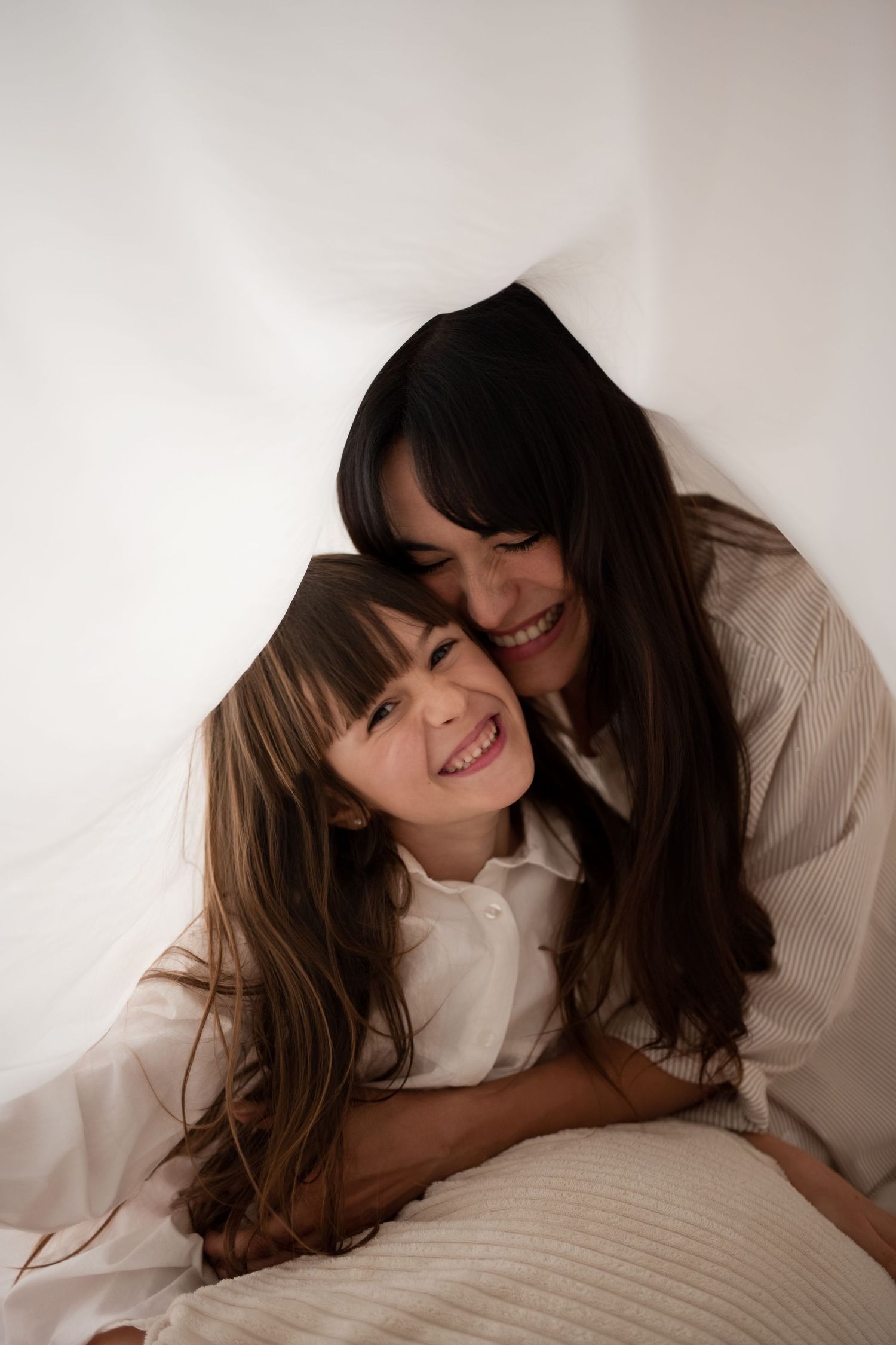 A mother with dark hair hugs her smiling daughter under a white blanket on a bed.