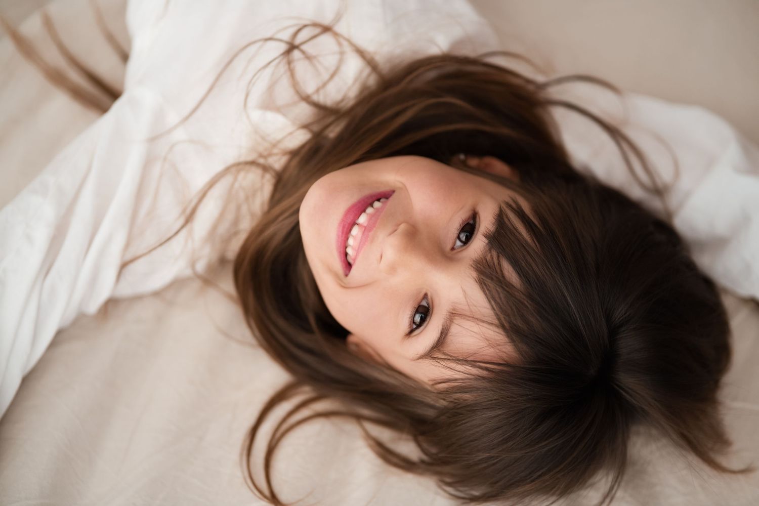 Young girl with dark hair lying back on white bedding smiling warmly up at camera.