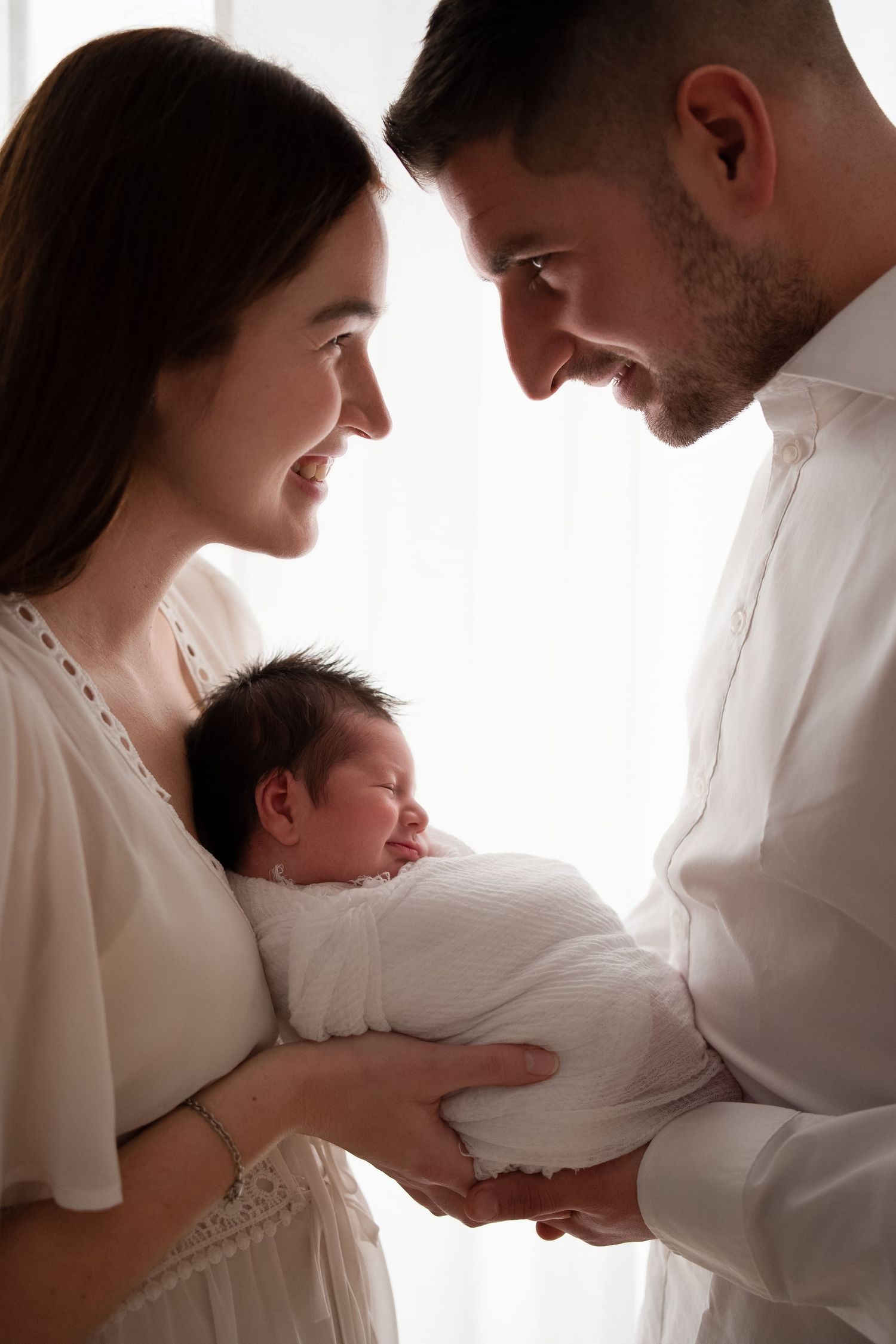 Happy parents smiling at their sleeping newborn baby wrapped in white cloth.