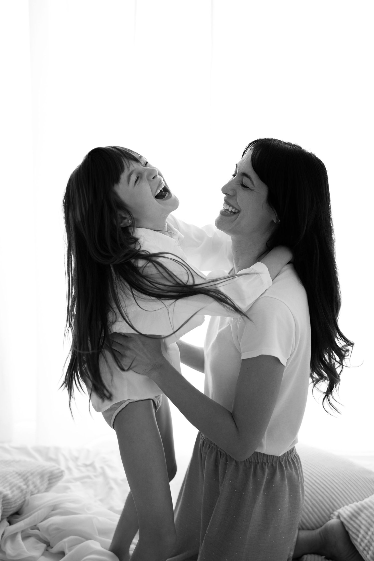 A laughing mother lifts her joyful daughter on a bed in a bright black and white photo.