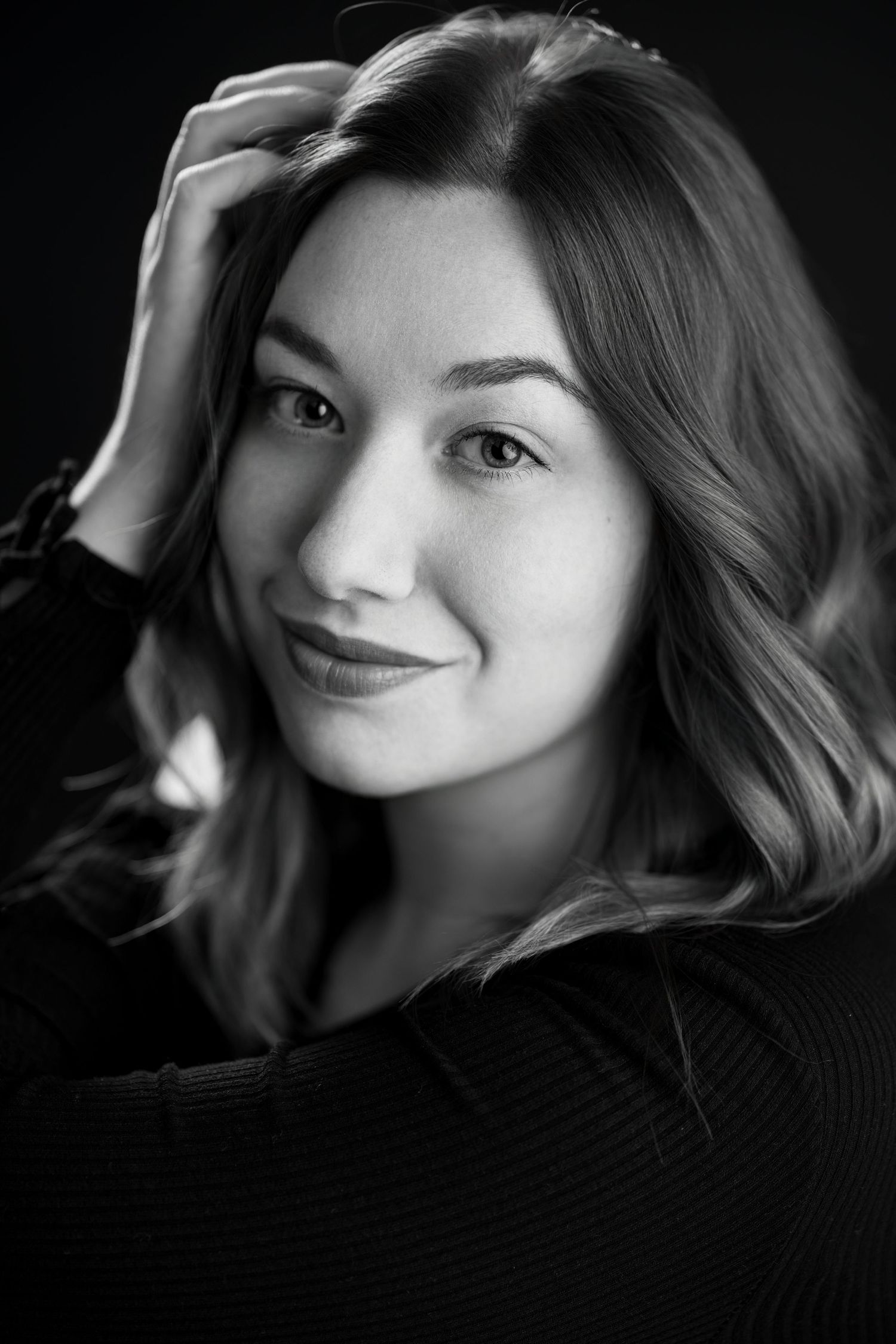 Black and white portrait of young woman with wavy hair smiling, hand resting on head.