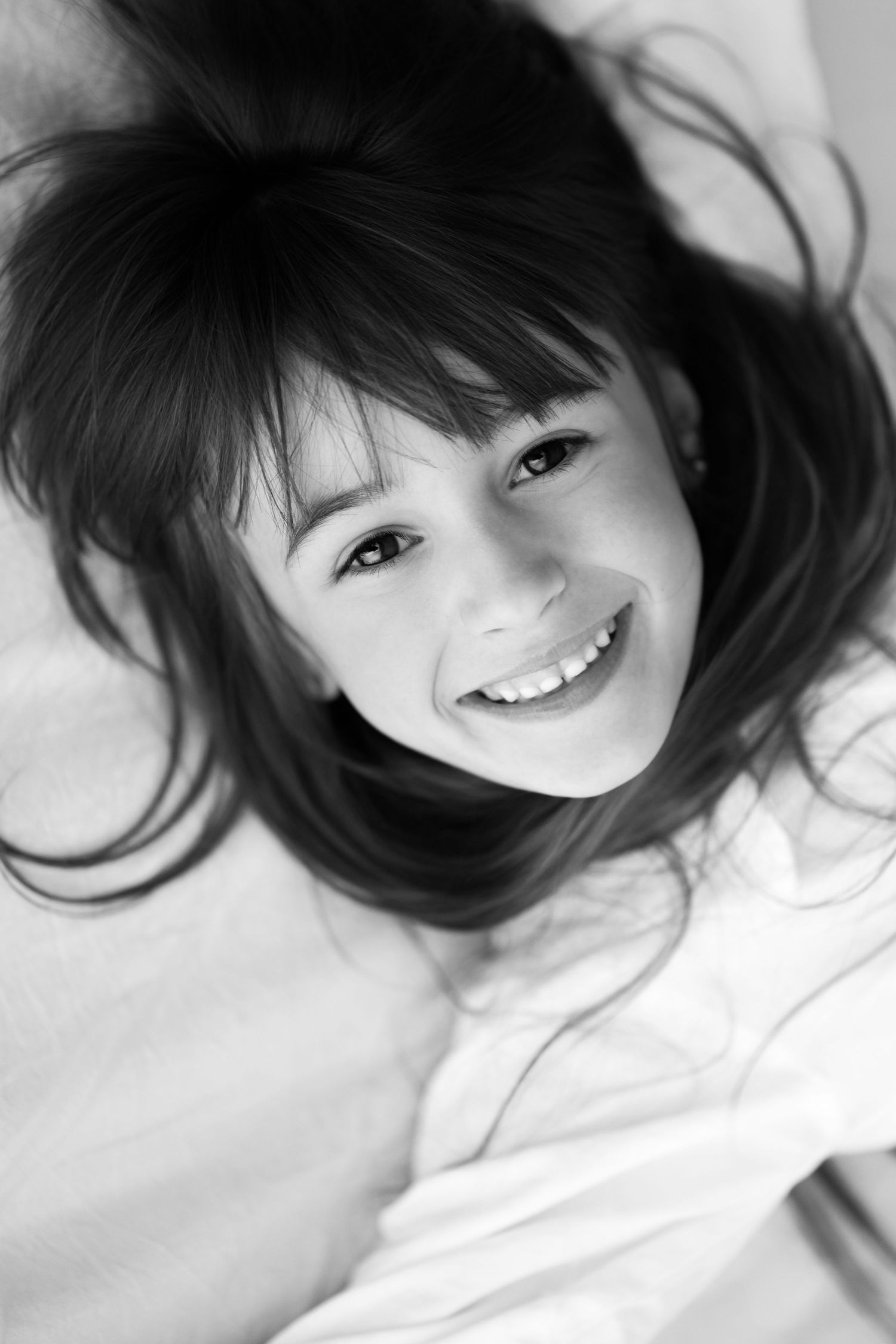 Black and white portrait of smiling young girl lying on white bed.