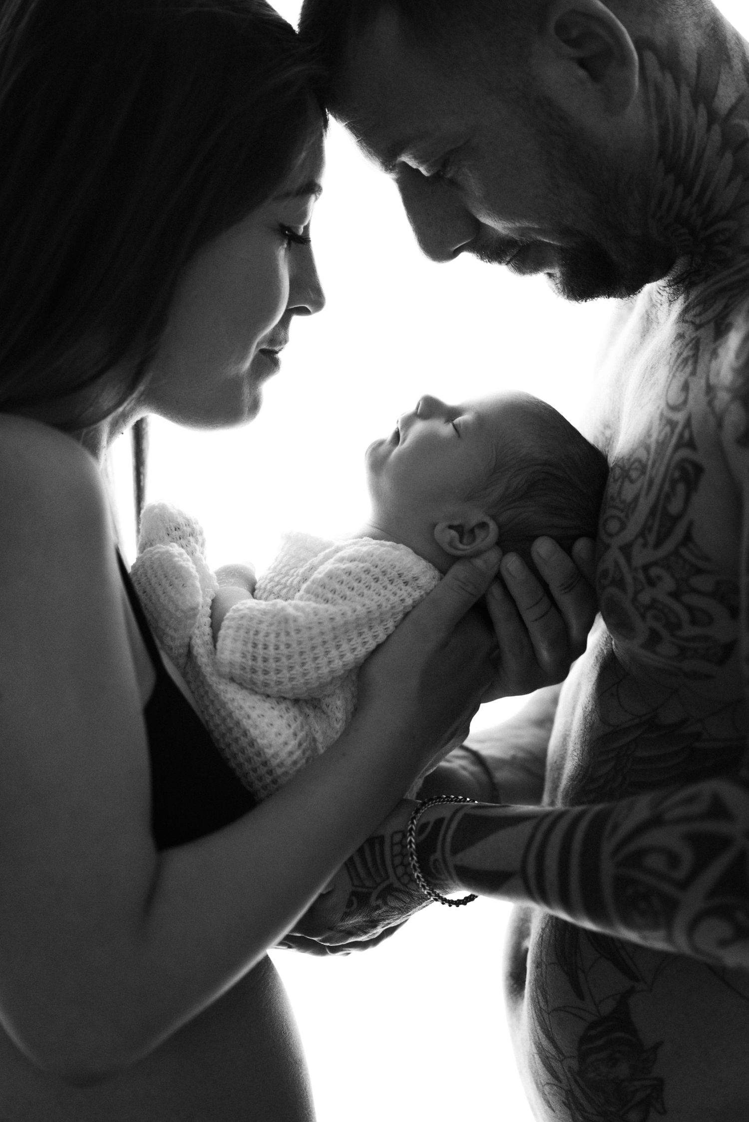 Black and white silhouette of tattooed father and mother tenderly holding their awake newborn baby.
