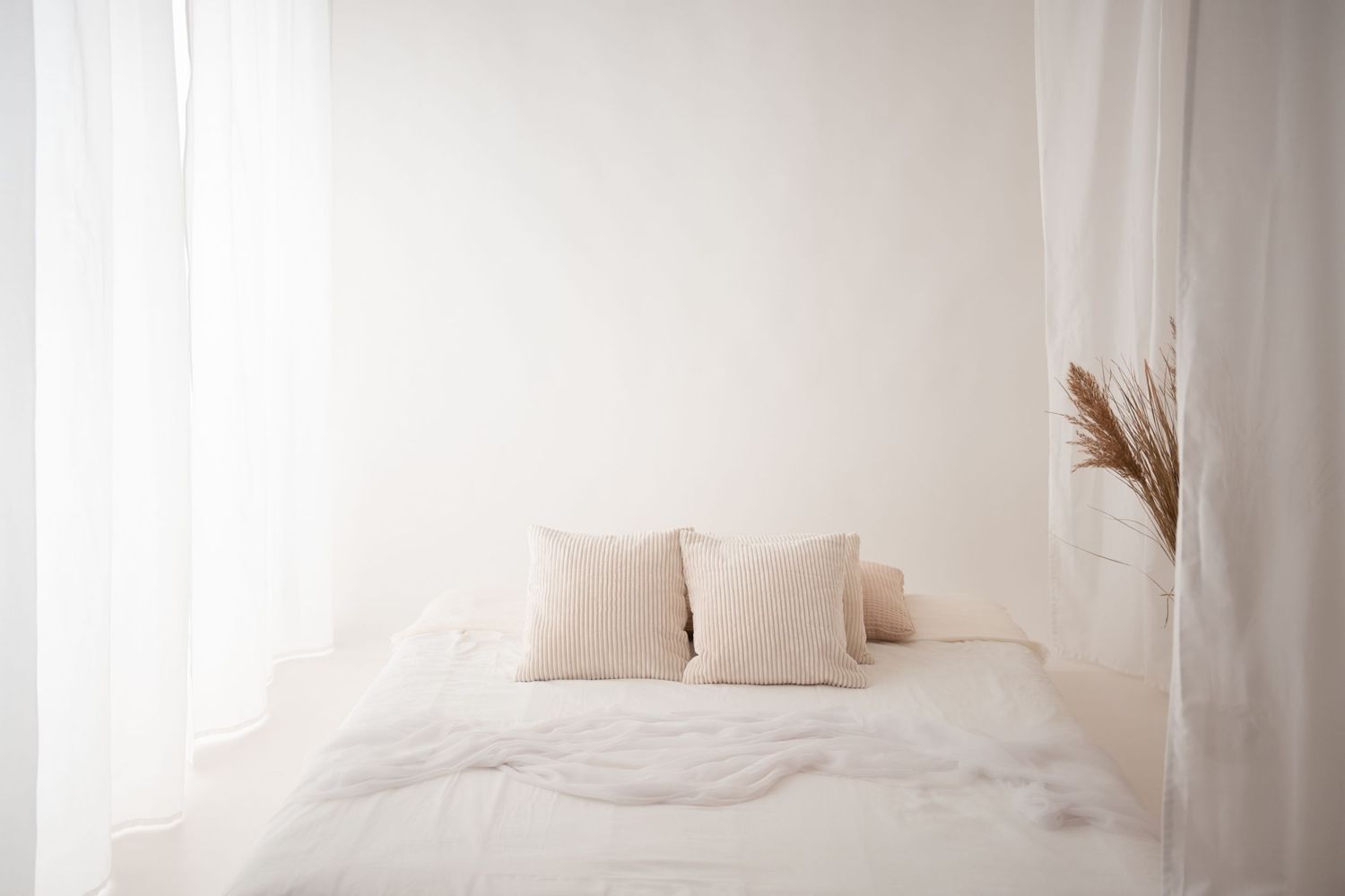 A minimalist white bedroom with a neatly made bed, beige pillows, and sheer white curtains.