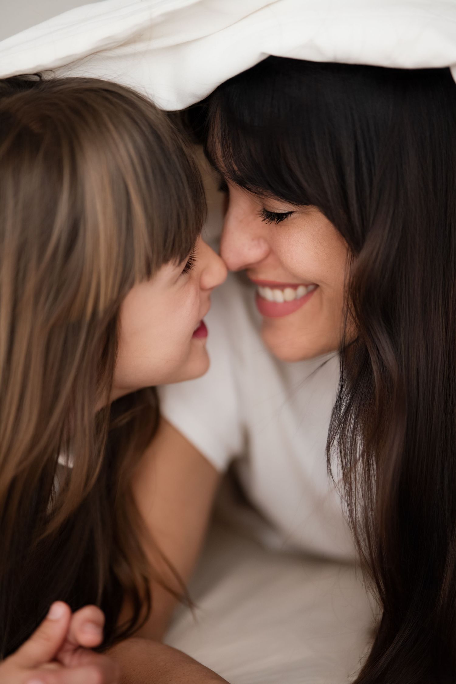 Mother and daughter smiling nose-to-nose under cozy white blanket on bed.