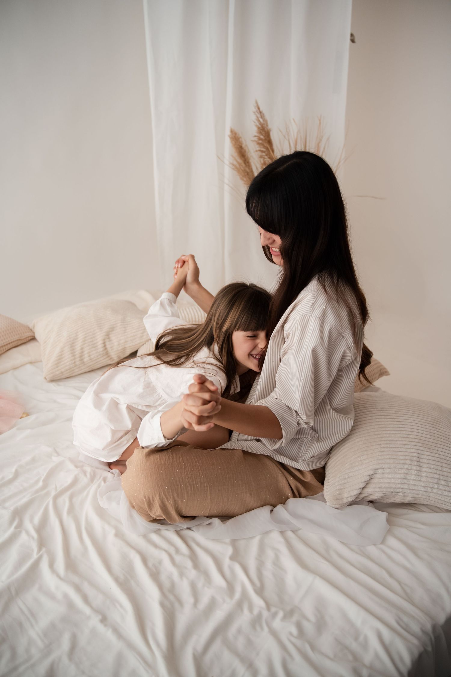A dark-haired mother sits cross-legged on a white bed, playfully holding hands with her young daughter.