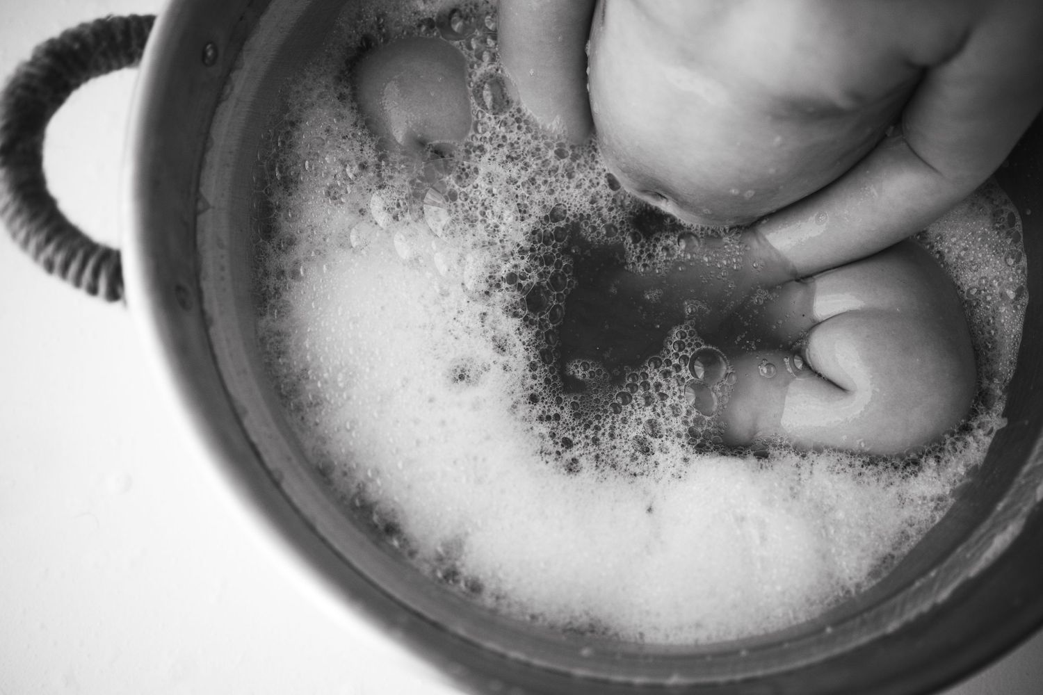 Close-up black and white photo of a baby's feet and hands in a soapy bubble bath in a metal tub.