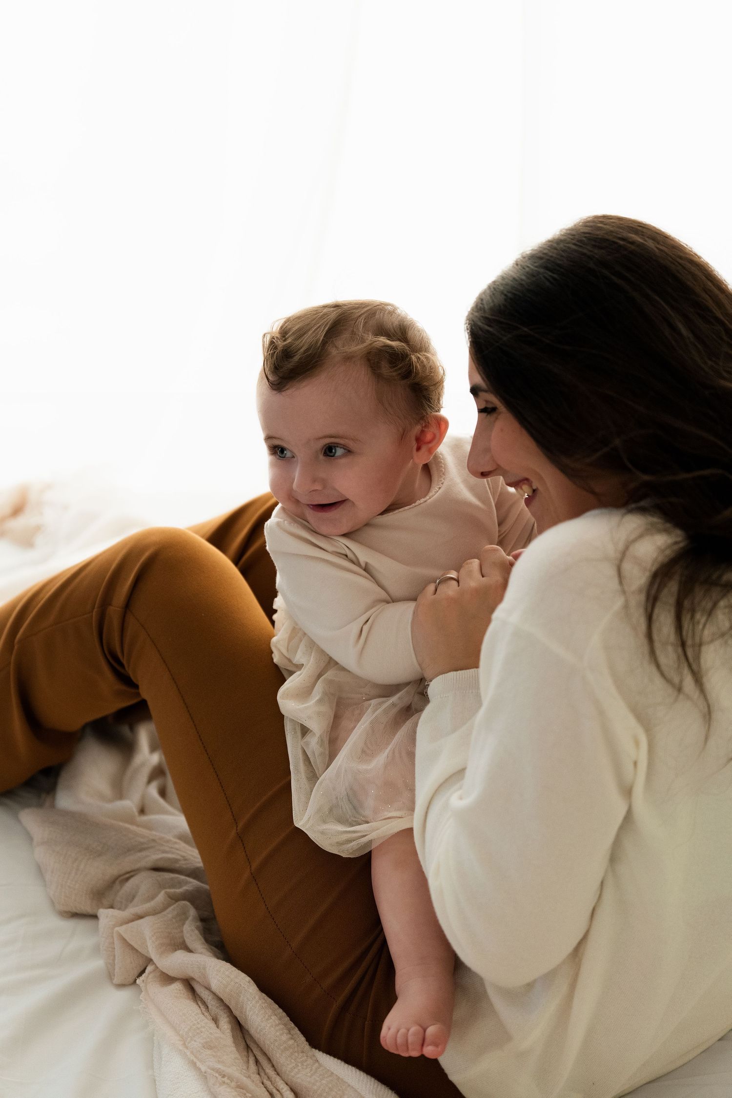 A smiling mother sits on a bed holding her happy baby in warm neutral tones.