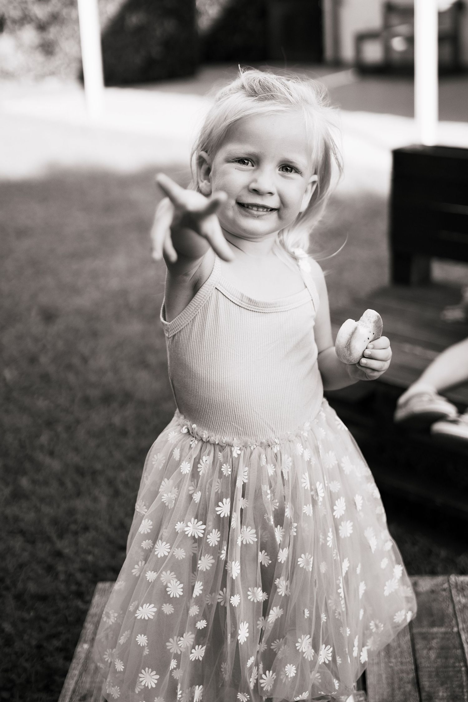 A cheerful little girl in a floral tutu dress reaches toward the camera outdoors.