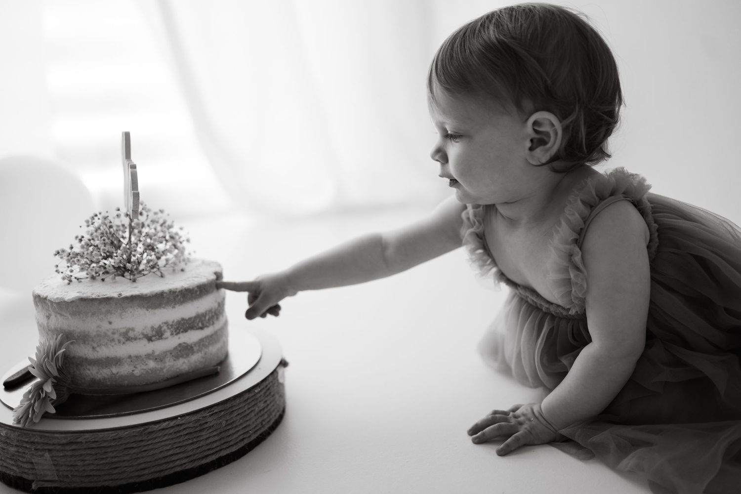 Black and white photo of a baby in a dress reaching toward a first birthday cake on a white table.