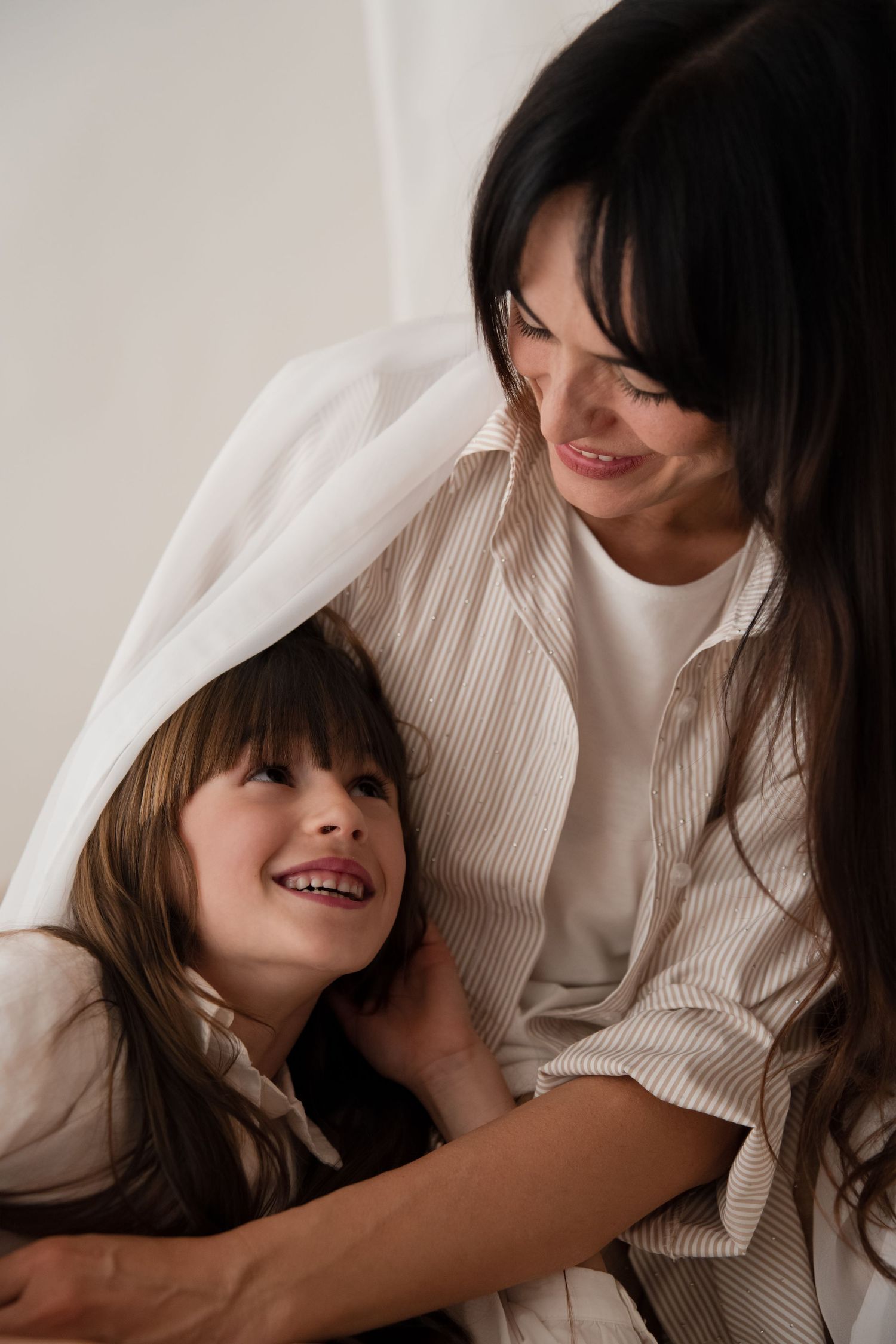Mother and daughter playing under white blanket, laughing together on bed.